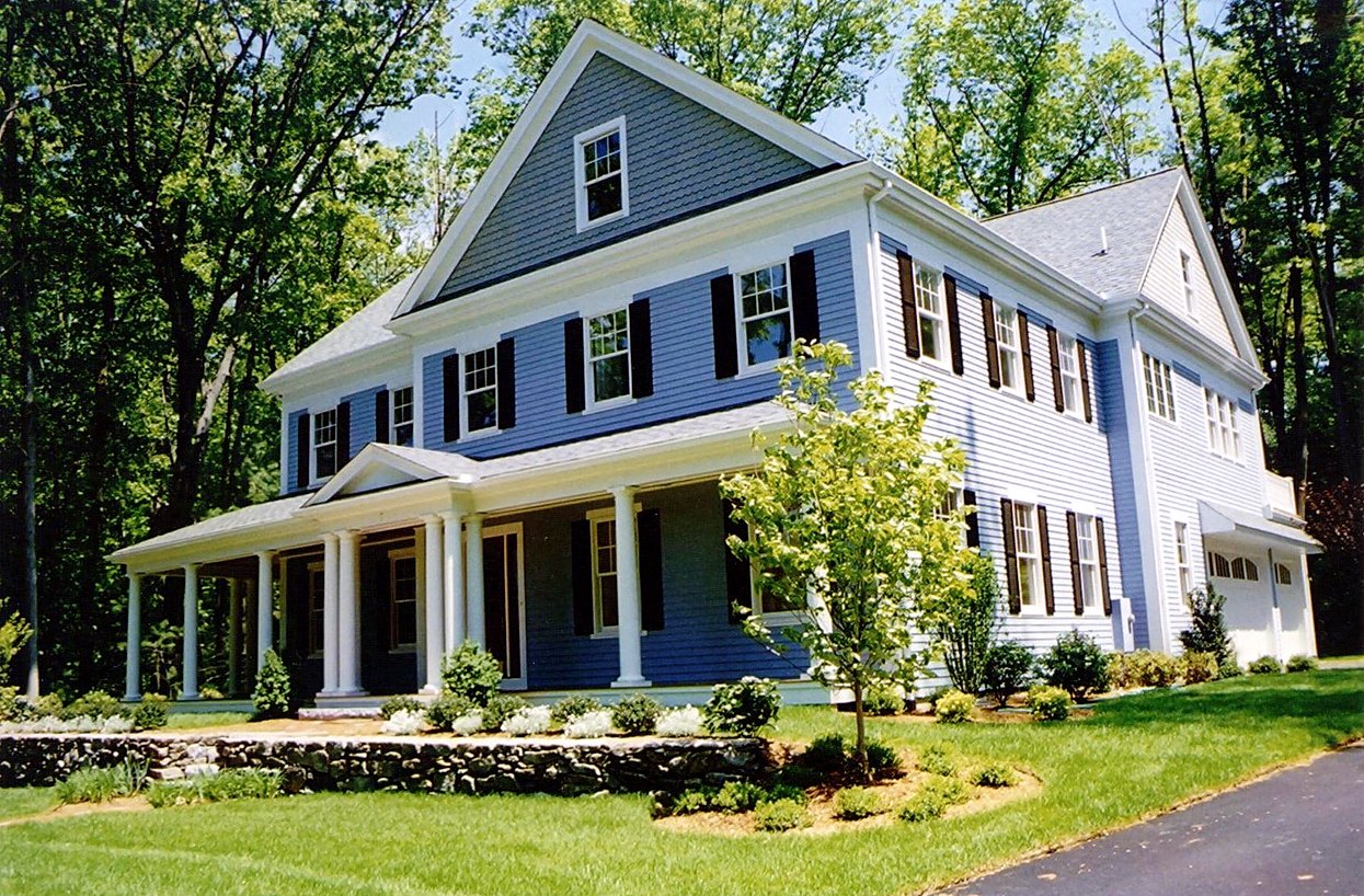 Blue colonial with a columned wraparound porch above a fieldstone wall in summer