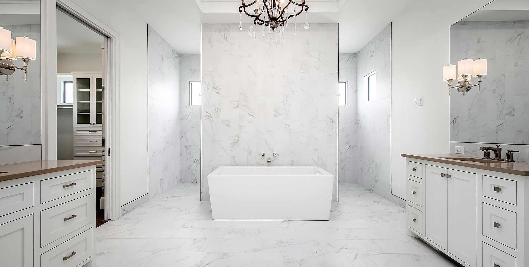 Primary bath with marble walls and floors, a freestanding tub and twin white vanities