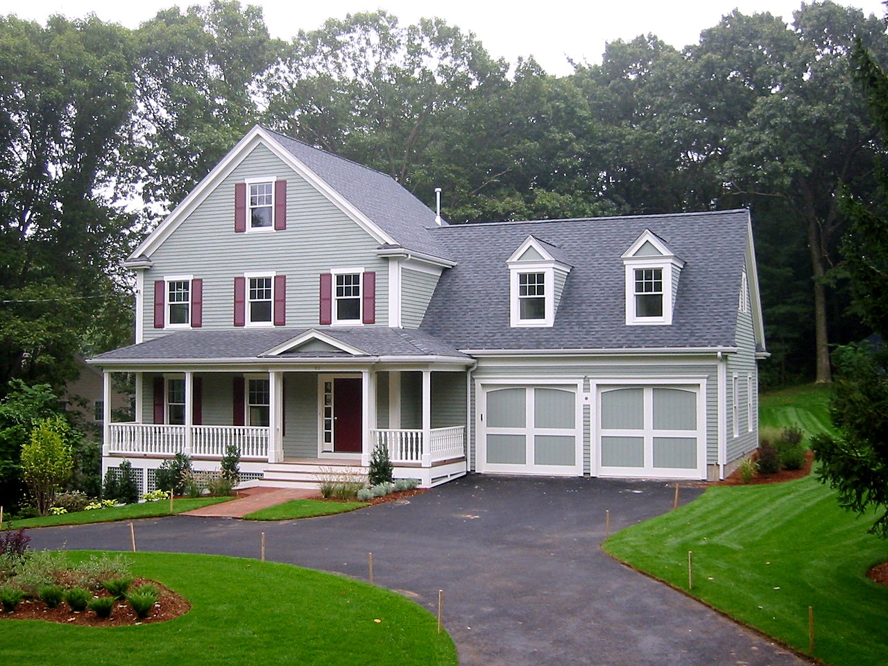 Sage-green farmhouse with a covered front porch, dormered garage wing and carriage doors